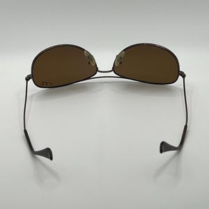 May include: Brown aviator sunglasses with a thin, dark metal frame. The lenses are a warm brown color, and the arms curve down to ear pieces. The sunglasses are set against a white background.