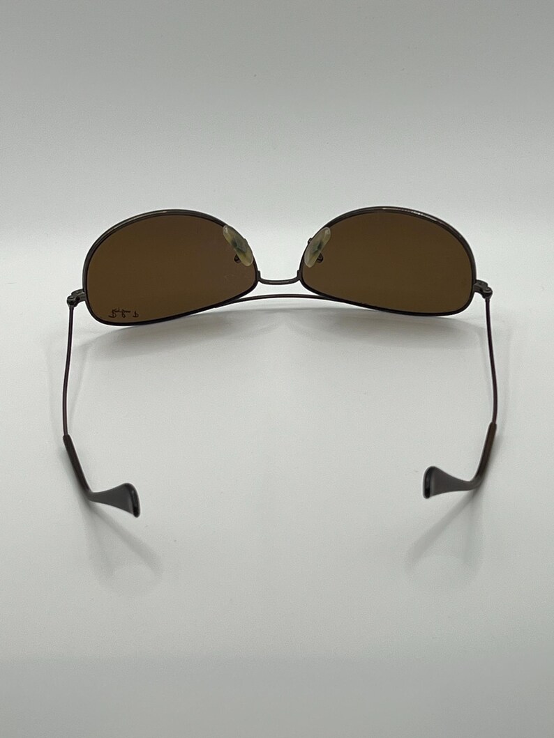 May include: Brown aviator sunglasses with a thin, dark metal frame. The lenses are a warm brown color, and the arms curve down to ear pieces. The sunglasses are set against a white background.