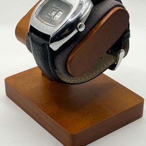 May include: A vintage digital watch with a black leather band and a silver-toned rectangular face. The watch displays the time and is mounted on a wooden display stand. The watch face has the word "Lucerne" printed on it.