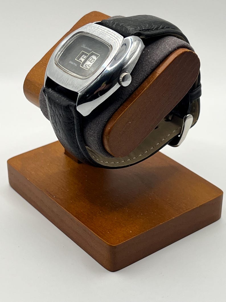 May include: A vintage digital watch with a black leather band and a silver-toned rectangular face. The watch displays the time and is mounted on a wooden display stand. The watch face has the word "Lucerne" printed on it.