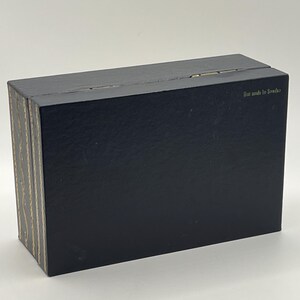 Puede incluir: Una caja negra rectangular con tapa con bisagras. El lateral de la caja tiene un patr&oacute;n decorativo dorado y beige. El texto "Box made in Sweden" est&aacute; impreso en la parte superior de la caja.