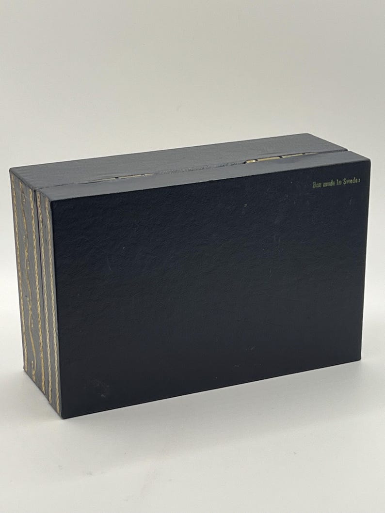 Puede incluir: Una caja negra rectangular con tapa con bisagras. El lateral de la caja tiene un patr&oacute;n decorativo dorado y beige. El texto "Box made in Sweden" est&aacute; impreso en la parte superior de la caja.