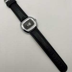 May include: A vintage wristwatch with a black leather band and a silver-toned, rectangular watch face. The watch face displays the time. The band is textured and has a silver buckle.
