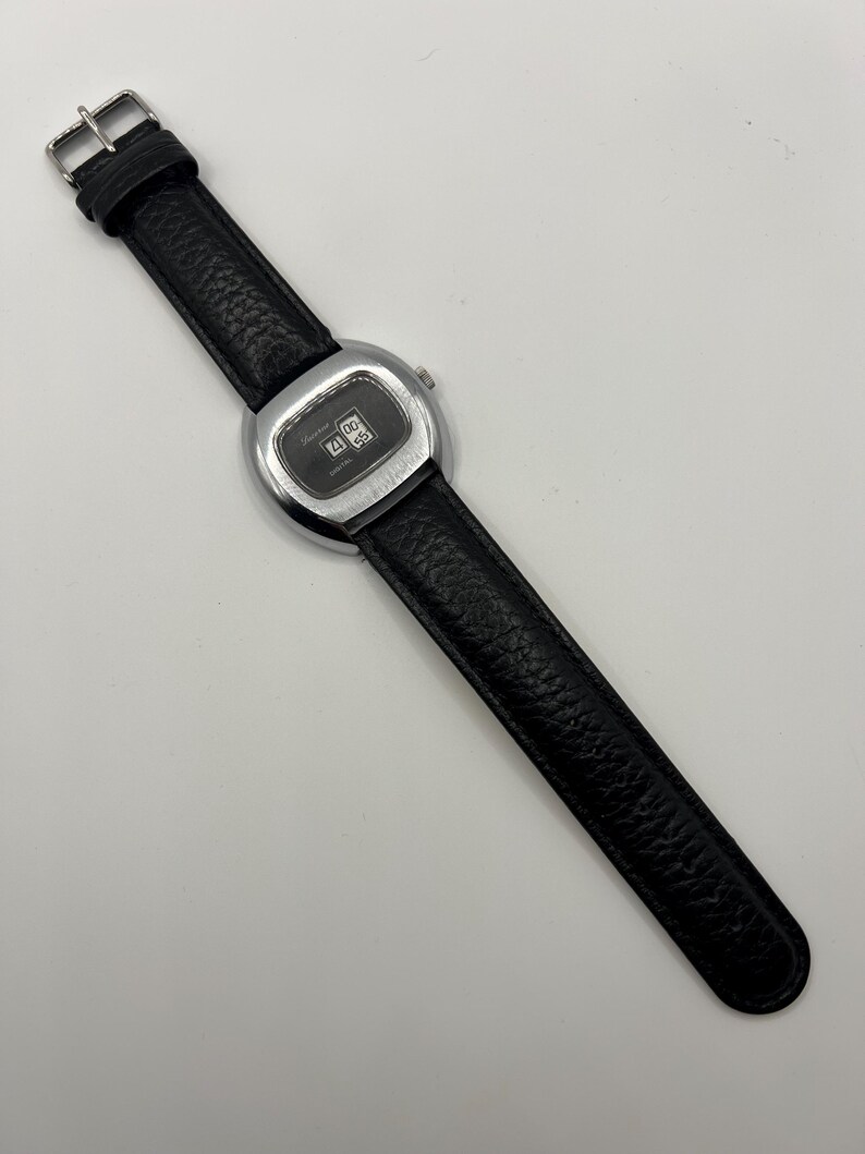 May include: A vintage wristwatch with a black leather band and a silver-toned, rectangular watch face. The watch face displays the time. The band is textured and has a silver buckle.