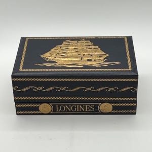 Puede incluir: Una caja rectangular negra con detalles dorados. La parte superior presenta una ilustraci&oacute;n dorada de un velero. La caja est&aacute; decorada con adornos dorados y la palabra "LONGINES" est&aacute; impresa en la parte delantera.