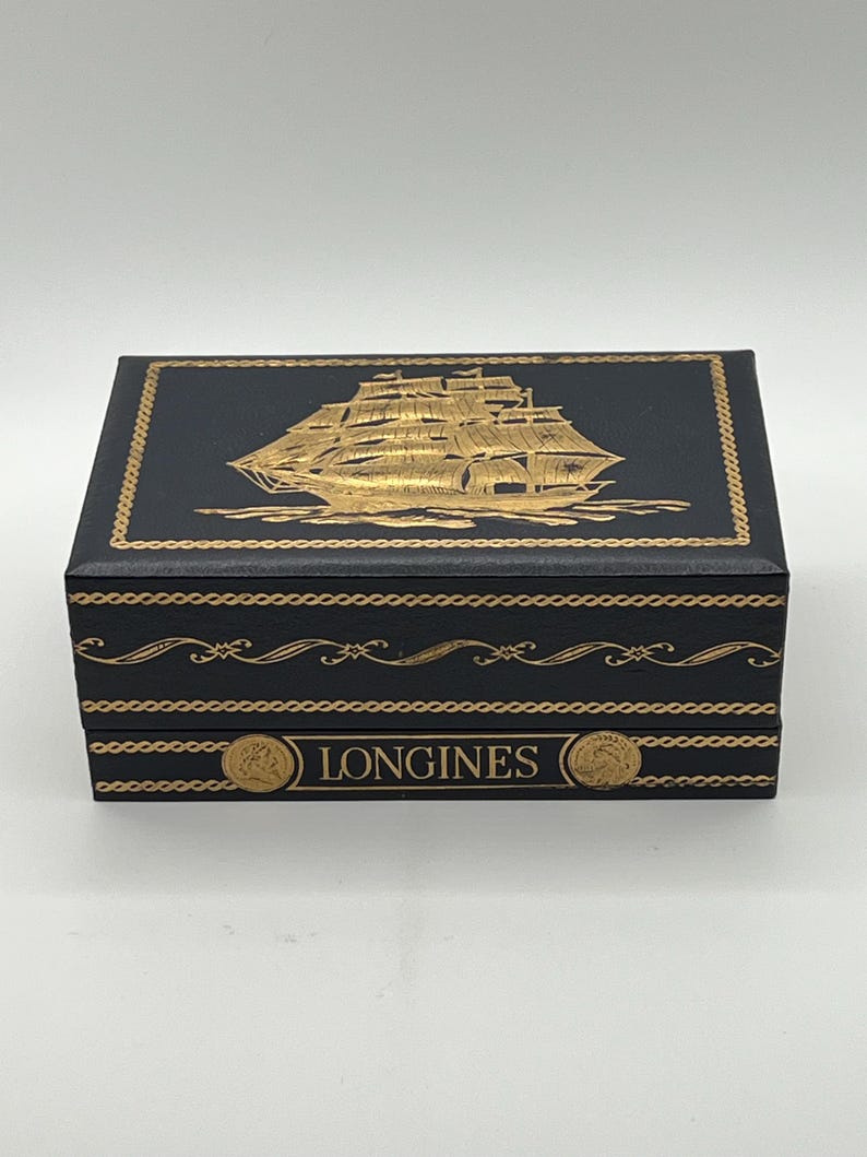 Puede incluir: Una caja rectangular negra con detalles dorados. La parte superior presenta una ilustraci&oacute;n dorada de un velero. La caja est&aacute; decorada con adornos dorados y la palabra "LONGINES" est&aacute; impresa en la parte delantera.