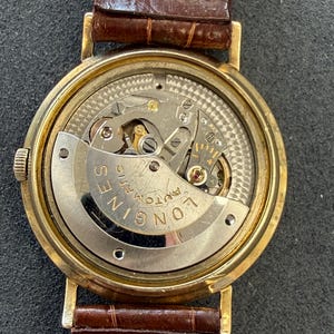 Puede incluir: Un reloj de pulsera autom&aacute;tico Longines dorado con una correa de cuero marr&oacute;n. El intrincado mecanismo interno del reloj es visible a trav&eacute;s de la parte trasera transparente. La esfera del reloj es redonda y la correa es de color marr&oacute;n intenso.
