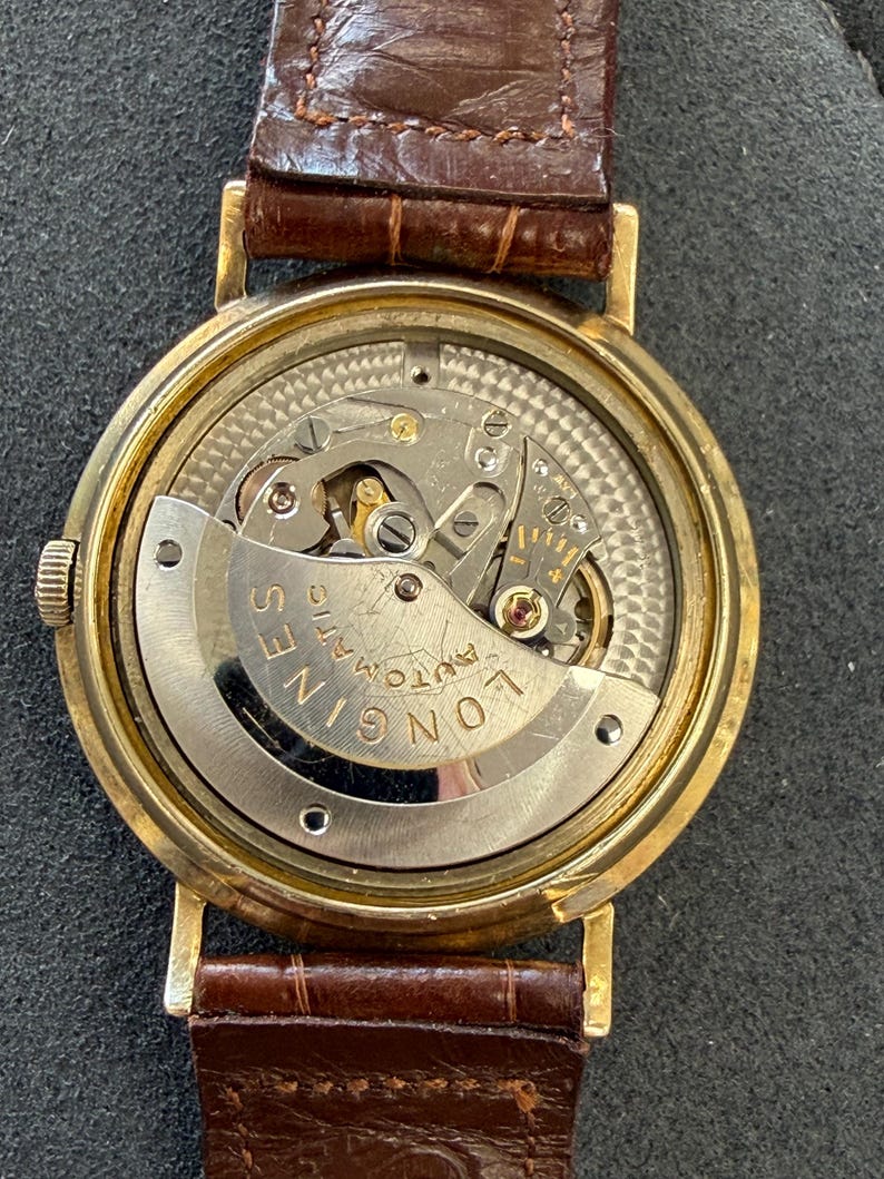 Puede incluir: Un reloj de pulsera autom&aacute;tico Longines dorado con una correa de cuero marr&oacute;n. El intrincado mecanismo interno del reloj es visible a trav&eacute;s de la parte trasera transparente. La esfera del reloj es redonda y la correa es de color marr&oacute;n intenso.