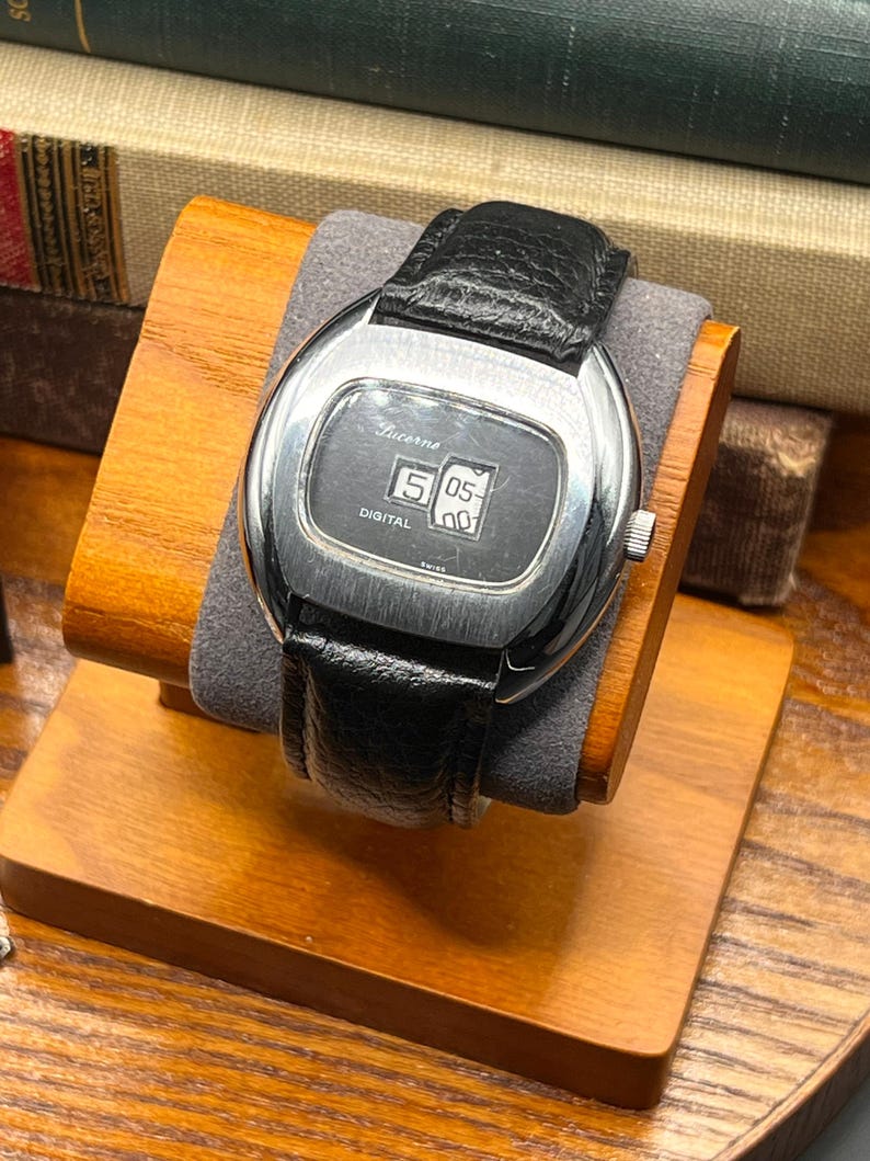 May include: A vintage Lucerne digital watch with a black leather band. The watch face is silver with a digital display showing the time. The watch is displayed on a wooden stand with a gray cushion.