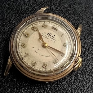 May include: Vintage Mido Multifort Grand Luxe watch with a gold-tone case and a cream-colored face. The watch features gold hands and hour markers, with the words "Super-Automatic" and "Mido Multifort Grand Luxe" visible on the face.