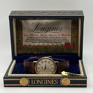 Puede incluir: Reloj Longines vintage en su caja de presentaci&oacute;n original. El reloj tiene una caja dorada, una esfera color crema y una correa de cuero marr&oacute;n. La caja es azul oscuro y negra con detalles dorados y el logotipo de Longines.