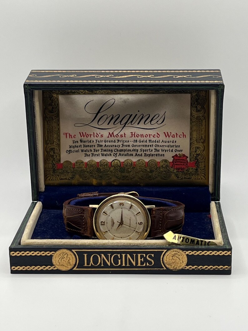 Puede incluir: Reloj Longines vintage en su caja de presentaci&oacute;n original. El reloj tiene una caja dorada, una esfera color crema y una correa de cuero marr&oacute;n. La caja es azul oscuro y negra con detalles dorados y el logotipo de Longines.