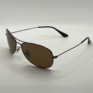 May include: Brown aviator sunglasses with a thin, dark metal frame. The lenses are a warm brown color, and the overall design is sleek and modern. The sunglasses are set against a plain white background.