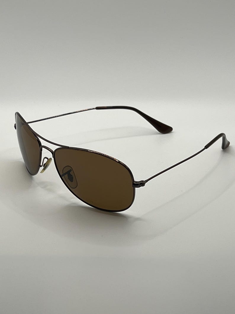 May include: Brown aviator sunglasses with a thin, dark metal frame. The lenses are a warm brown color, and the overall design is sleek and modern. The sunglasses are set against a plain white background.