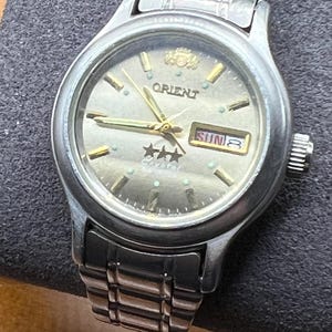 May include: A silver-toned Orient wristwatch with a gray face, gold hands, and hour markers. The watch displays the day and date, with the day reading "SUN 8". The watch has a silver band and a textured bezel.