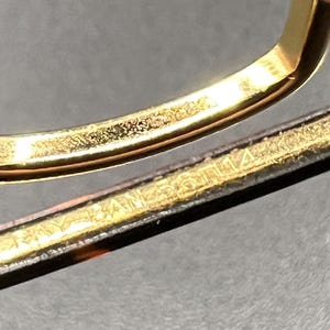 May include: Close-up of a gold-colored metal frame, possibly for eyeglasses or sunglasses. The frame has a shiny, reflective surface. The text "RAY BAN" is visible on the frame.