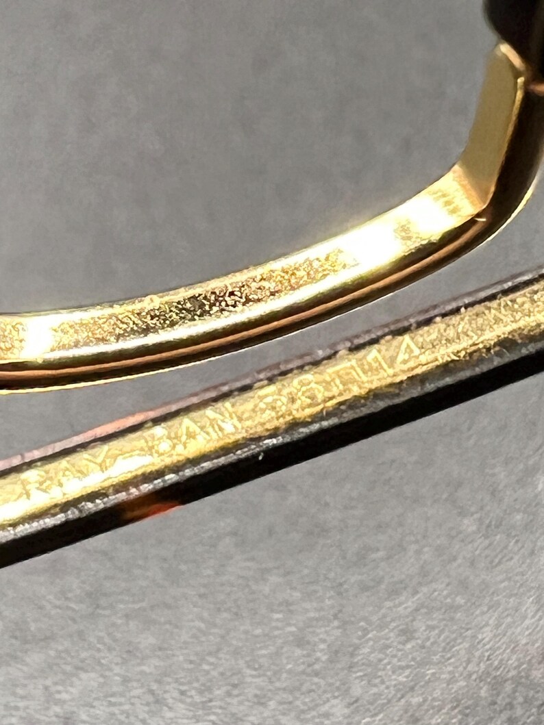 May include: Close-up of a gold-colored metal frame, possibly for eyeglasses or sunglasses. The frame has a shiny, reflective surface. The text "RAY BAN" is visible on the frame.