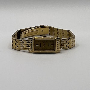 May include: A gold-toned wristwatch with a rectangular face and a matching link band. The watch has a simple, elegant design, with a gold dial and hour markers. The band is made of small, rectangular links that connect to form a flexible bracelet.