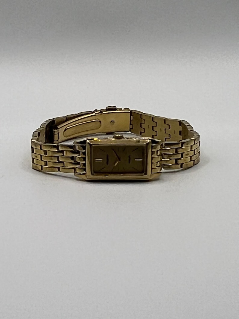 May include: A gold-toned wristwatch with a rectangular face and a matching link band. The watch has a simple, elegant design, with a gold dial and hour markers. The band is made of small, rectangular links that connect to form a flexible bracelet.