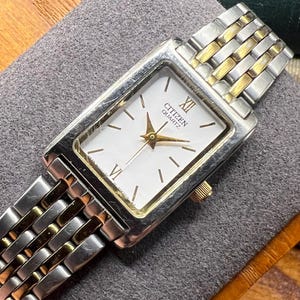 May include: A Citizen Quartz watch with a rectangular white face, gold hands, and gold and silver band. The watch face displays the Citizen Quartz logo and Roman numerals. The watch is a classic design.