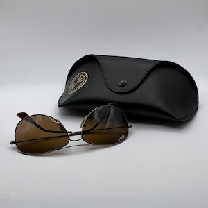May include: Brown aviator sunglasses with a black case. The sunglasses have a thin, dark metal frame and brown-tinted lenses. The black case has a button closure and a gold emblem.