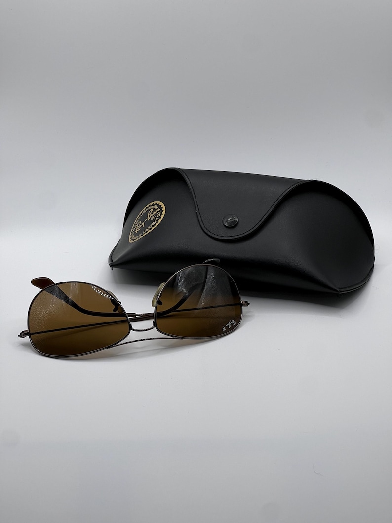 May include: Brown aviator sunglasses with a black case. The sunglasses have a thin, dark metal frame and brown-tinted lenses. The black case has a button closure and a gold emblem.