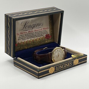 Puede incluir: Un reloj Longines vintage en su caja original. El reloj tiene una caja dorada, una esfera clara y una correa de cuero marr&oacute;n. La caja es negra con detalles dorados y el logotipo de Longines.