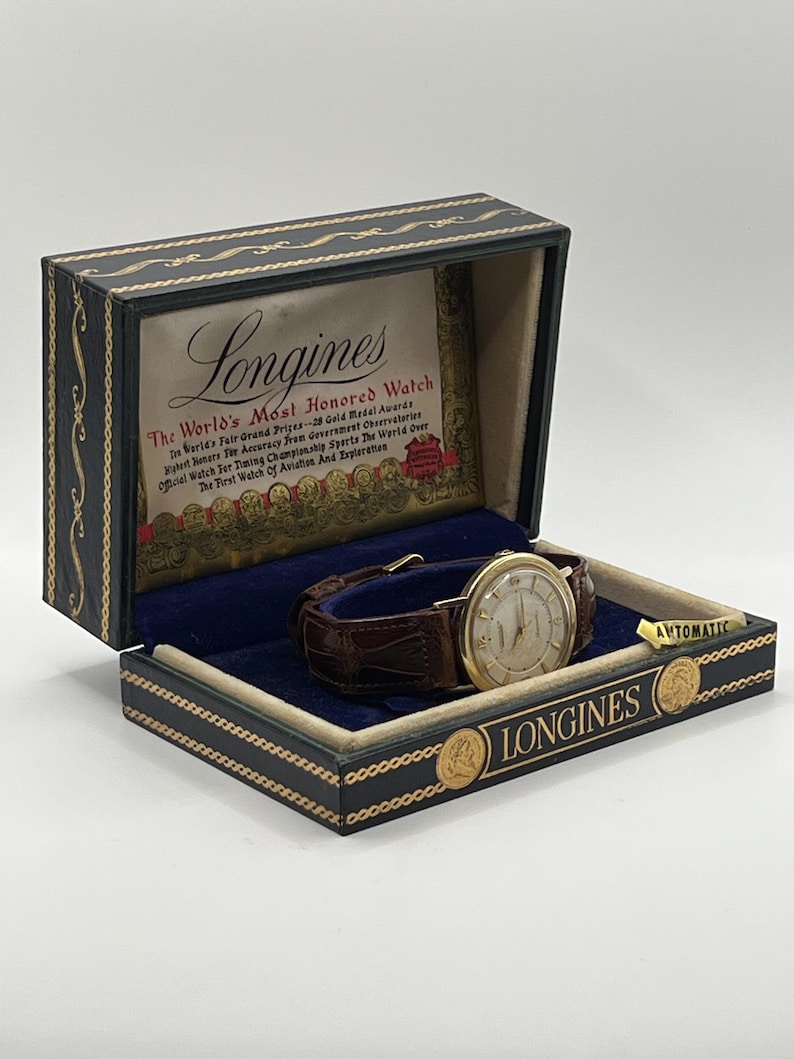 Puede incluir: Un reloj Longines vintage en su caja original. El reloj tiene una caja dorada, una esfera clara y una correa de cuero marr&oacute;n. La caja es negra con detalles dorados y el logotipo de Longines.