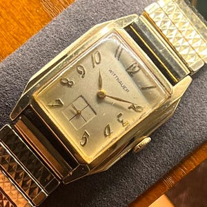 May include: A vintage gold-toned Wittnauer wristwatch with a rectangular face. The watch features gold hands and hour markers, with the brand name displayed. The watch band is a gold-toned, expandable style.