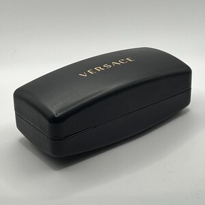 Peut inclure: Un &eacute;tui &agrave; lunettes rectangulaire noir avec le mot "VERSACE" dor&eacute; imprim&eacute; sur le dessus. L'&eacute;tui est ferm&eacute; et pr&eacute;sente une finition lisse et mate. L'&eacute;tui est probablement en cuir ou dans un mat&eacute;riau similaire.
