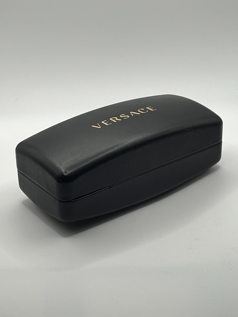 Peut inclure: Un &eacute;tui &agrave; lunettes rectangulaire noir avec le mot "VERSACE" dor&eacute; imprim&eacute; sur le dessus. L'&eacute;tui est ferm&eacute; et pr&eacute;sente une finition lisse et mate. L'&eacute;tui est probablement en cuir ou dans un mat&eacute;riau similaire.