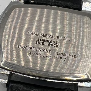 May include: Close-up of a vintage watch back with a black leather strap. The watch back is silver with the text "BASE METAL BEZEL, STAINLESS STEEL BACK, SHOCKRESISTANT, SWISS MADE, 12712-1 1957".
