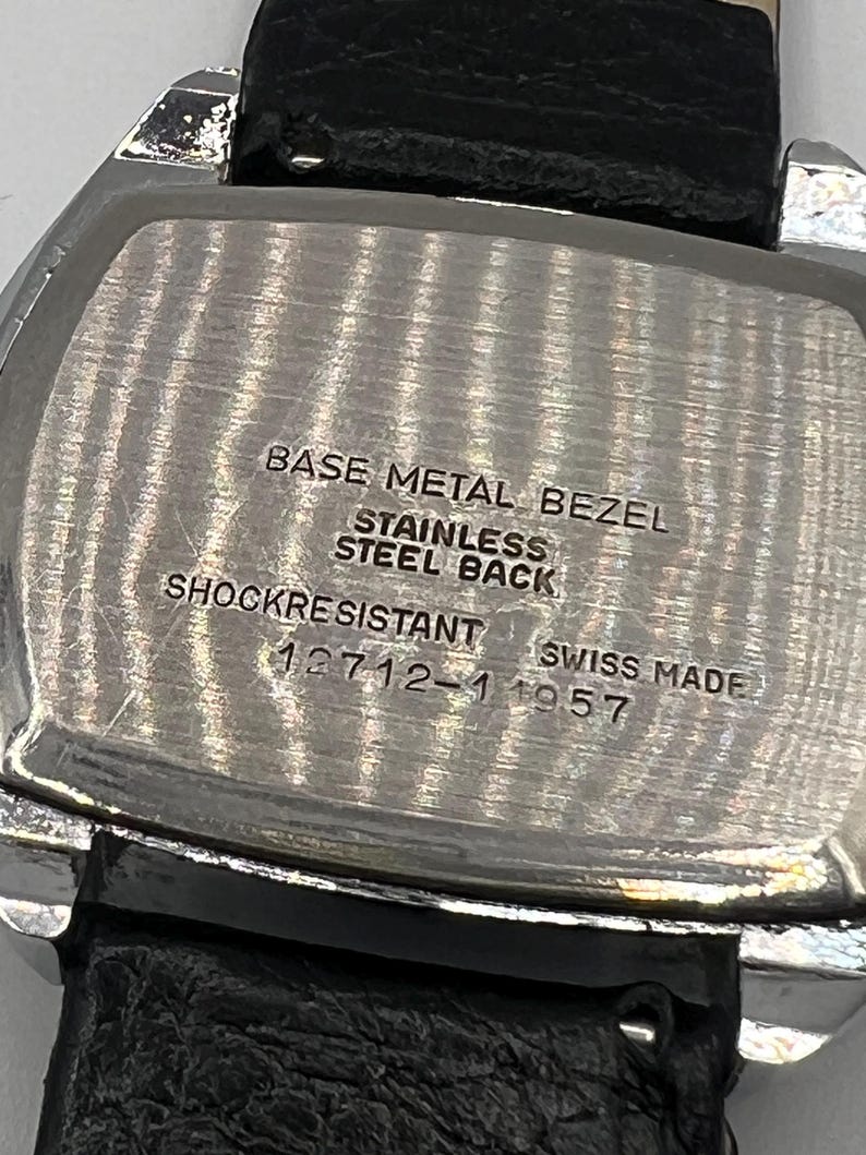 May include: Close-up of a vintage watch back with a black leather strap. The watch back is silver with the text "BASE METAL BEZEL, STAINLESS STEEL BACK, SHOCKRESISTANT, SWISS MADE, 12712-1 1957".