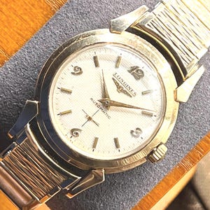 May include: A vintage gold-toned Longines automatic wristwatch with a white face. The watch features gold hands and hour markers, a subdial, and the word "AUTOMATIC" is printed on the face. The watch has a gold-toned band.