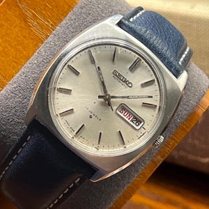 May include: A vintage Seiko automatic watch with a silver dial and a blue leather strap. The watch face displays the day and date, with the day reading "SUN 26". The watch has a stainless steel case and is marked "17 JEWELS".