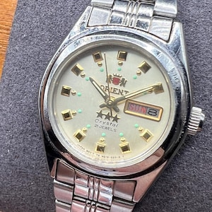 May include: Silver-toned Orient Crystal watch with a cream-colored face. The watch features gold-colored hour markers and hands, with a day-date display at the 3 o'clock position. The watch band is made of silver-toned metal links.