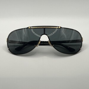 Peut inclure: Lunettes de soleil noires en forme d'&eacute;cran avec des verres fonc&eacute;s et des d&eacute;tails dor&eacute;s. Ces lunettes pr&eacute;sentent un design moderne avec un pont et une barre sup&eacute;rieure. Les verres sont gris fonc&eacute; et la monture est noire.