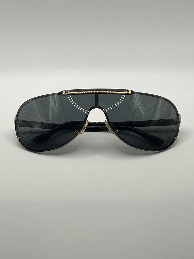 Peut inclure: Lunettes de soleil noires en forme d'&eacute;cran avec des verres fonc&eacute;s et des d&eacute;tails dor&eacute;s. Ces lunettes pr&eacute;sentent un design moderne avec un pont et une barre sup&eacute;rieure. Les verres sont gris fonc&eacute; et la monture est noire.