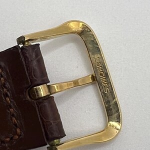 Puede incluir: Primer plano de una hebilla de reloj dorada con el nombre de la marca "LONGINES" grabado. La hebilla est&aacute; sujeta a una correa de reloj de cuero marr&oacute;n con costuras visibles.