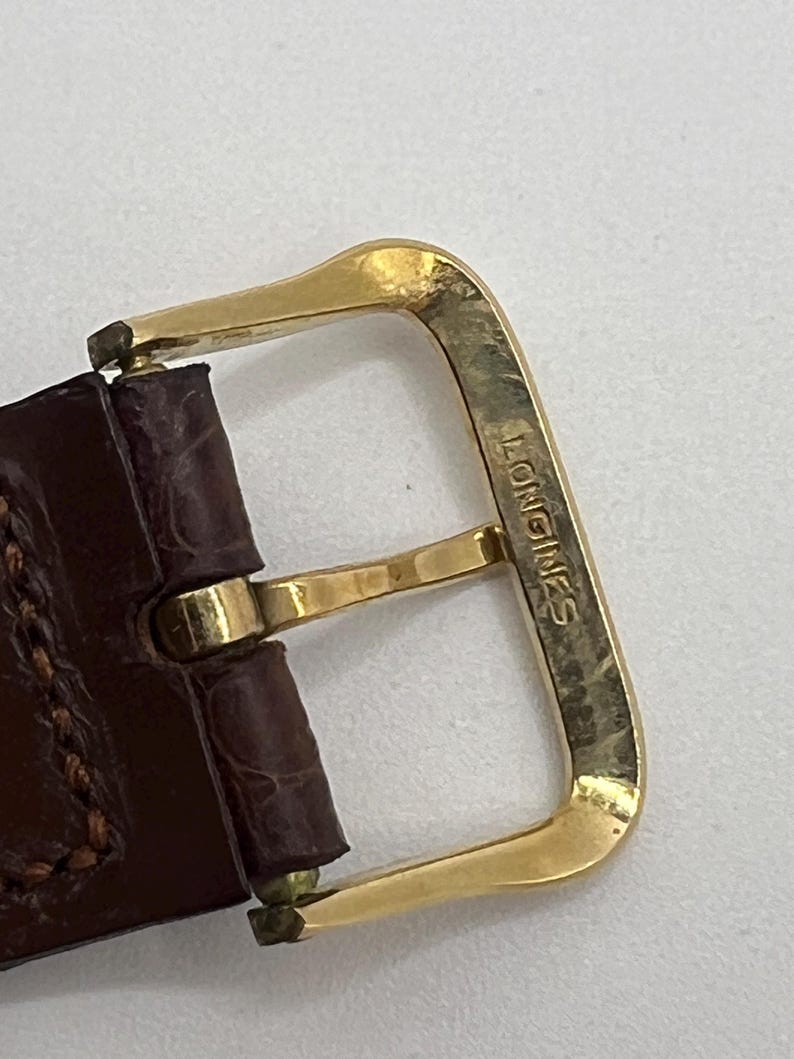 Puede incluir: Primer plano de una hebilla de reloj dorada con el nombre de la marca "LONGINES" grabado. La hebilla est&aacute; sujeta a una correa de reloj de cuero marr&oacute;n con costuras visibles.