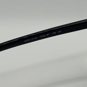 Peut inclure: Gros plan sur la branche d'une paire de lunettes noires. Le texte "MOD. 2140 1002/87 135 3N" est imprim&eacute; sur la branche. Les lunettes sont pos&eacute;es sur un fond blanc.