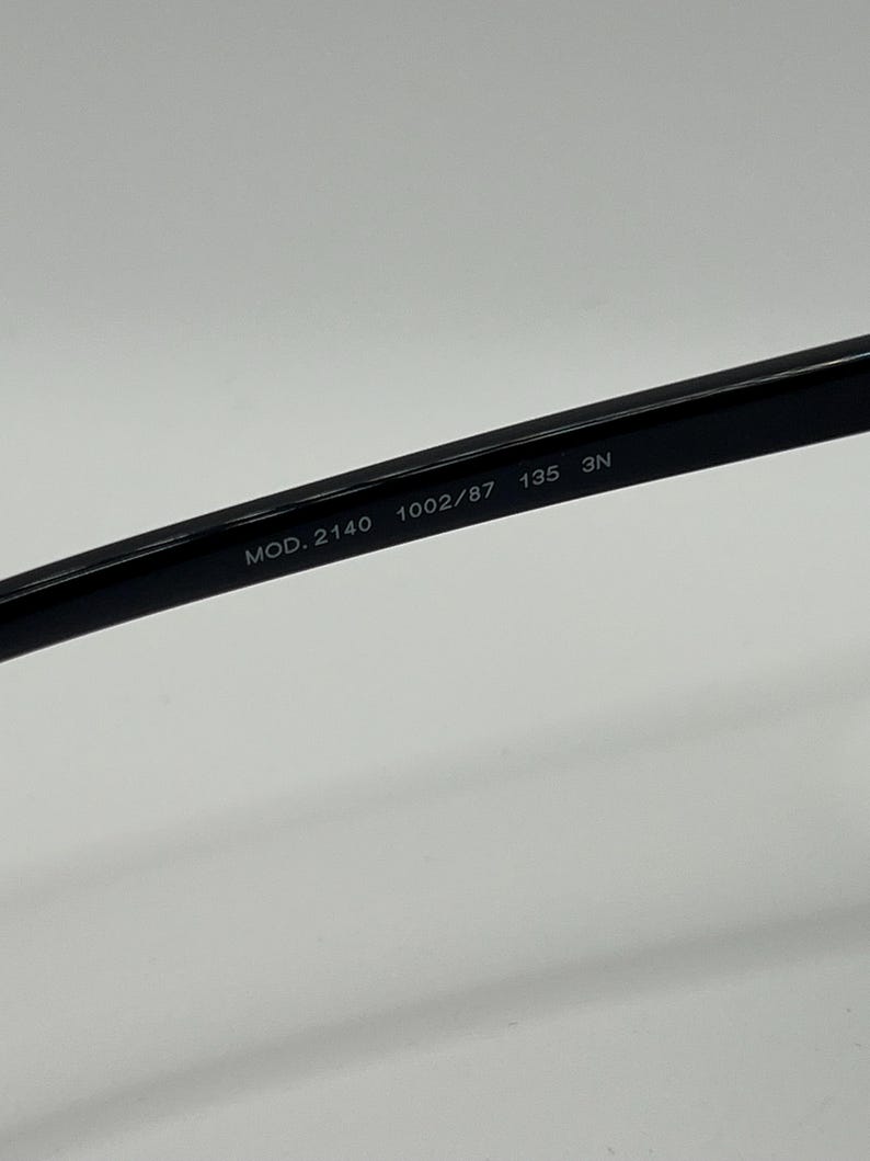 Peut inclure: Gros plan sur la branche d'une paire de lunettes noires. Le texte "MOD. 2140 1002/87 135 3N" est imprim&eacute; sur la branche. Les lunettes sont pos&eacute;es sur un fond blanc.