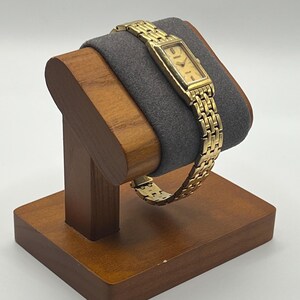 May include: A gold-toned wristwatch with a rectangular face and a linked band is displayed on a wooden watch stand. The watch face has a light-colored background with black hour markers and hands. The stand is made of dark wood and has a gray cushion.