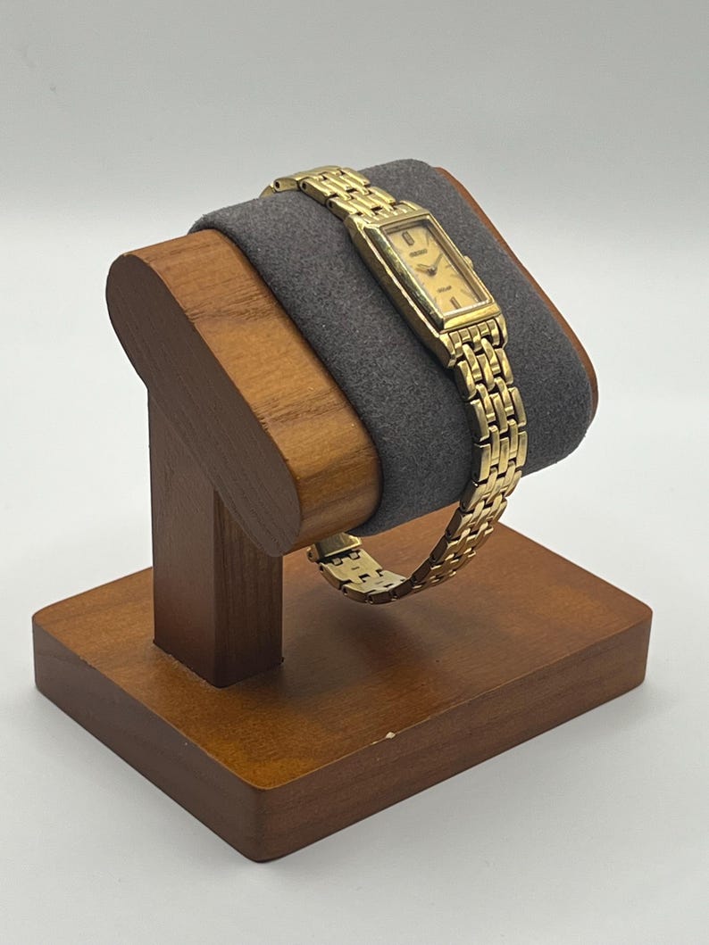 May include: A gold-toned wristwatch with a rectangular face and a linked band is displayed on a wooden watch stand. The watch face has a light-colored background with black hour markers and hands. The stand is made of dark wood and has a gray cushion.