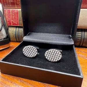 May include: A pair of oval cufflinks with a black and white houndstooth pattern, presented in a black box with a satin lining. The cufflinks have a silver-tone metal border. The box is open, revealing the cufflinks.