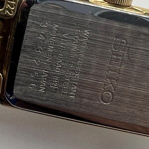 May include: A gold-toned rectangular Seiko watch with a silver-colored back. The back of the watch has the text "WATER RESISTANT STAINLESS STEEL" and "MOVEMENT JAPAN". The watch has a gold-colored crown and a gold-colored band.