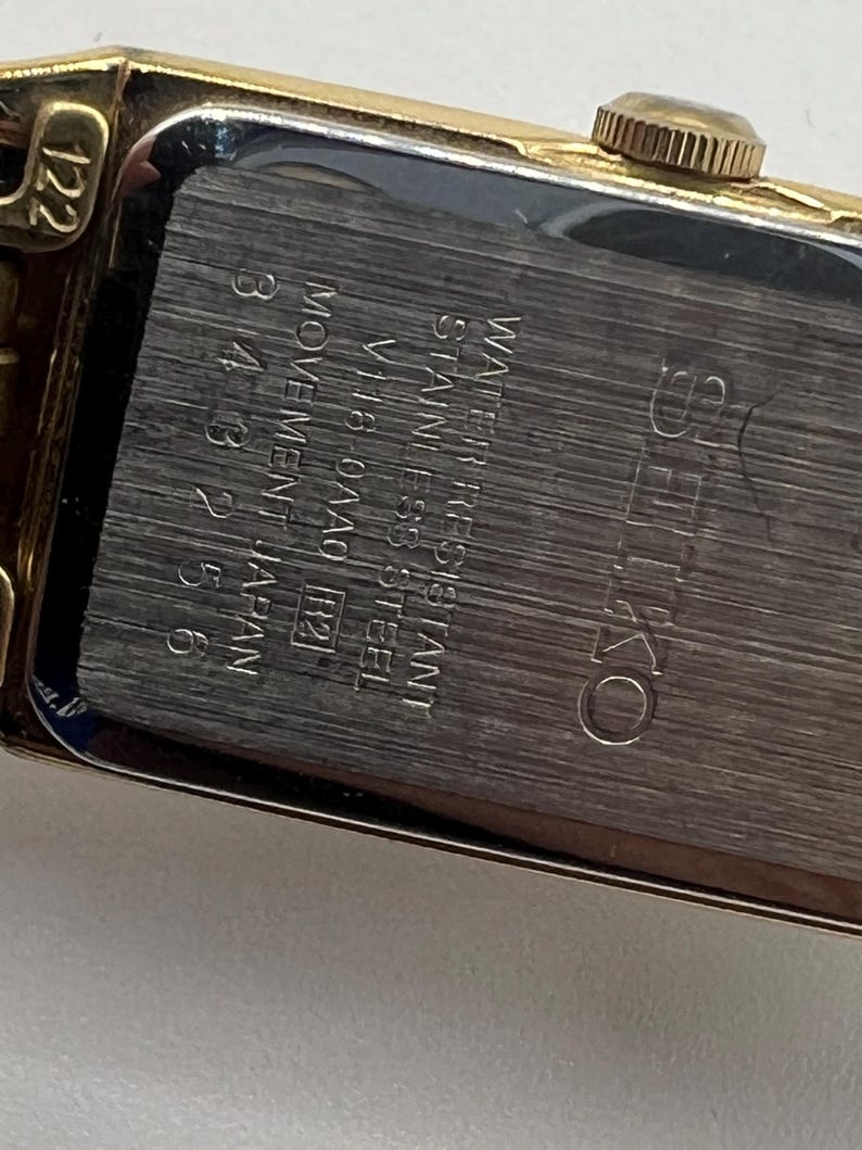 May include: A gold-toned rectangular Seiko watch with a silver-colored back. The back of the watch has the text "WATER RESISTANT STAINLESS STEEL" and "MOVEMENT JAPAN". The watch has a gold-colored crown and a gold-colored band.