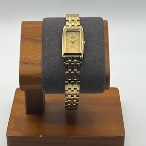 May include: A gold-toned Seiko Solar watch with a rectangular face and a matching link bracelet. The watch is displayed on a wooden watch stand with a gray velvet cushion. The watch face has gold hands and markers.