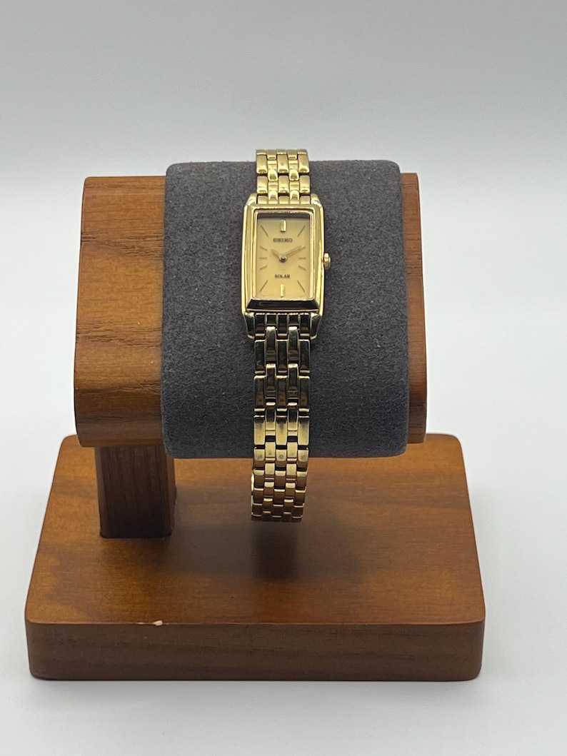 May include: A gold-toned Seiko Solar watch with a rectangular face and a matching link bracelet. The watch is displayed on a wooden watch stand with a gray velvet cushion. The watch face has gold hands and markers.