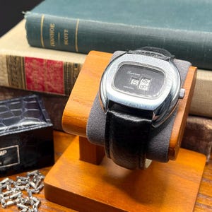May include: A vintage Lucerne digital watch with a black leather band, displayed on a wooden stand. The watch face shows the time as 5:00. The watch has a silver-colored case and is set against a backdrop of old books.